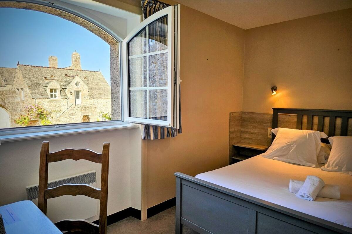 Une chambre à coucher avec un lit, une chaise, une fenêtre avec des rideaux et une vue sur un bâtiment à l’extérieur.