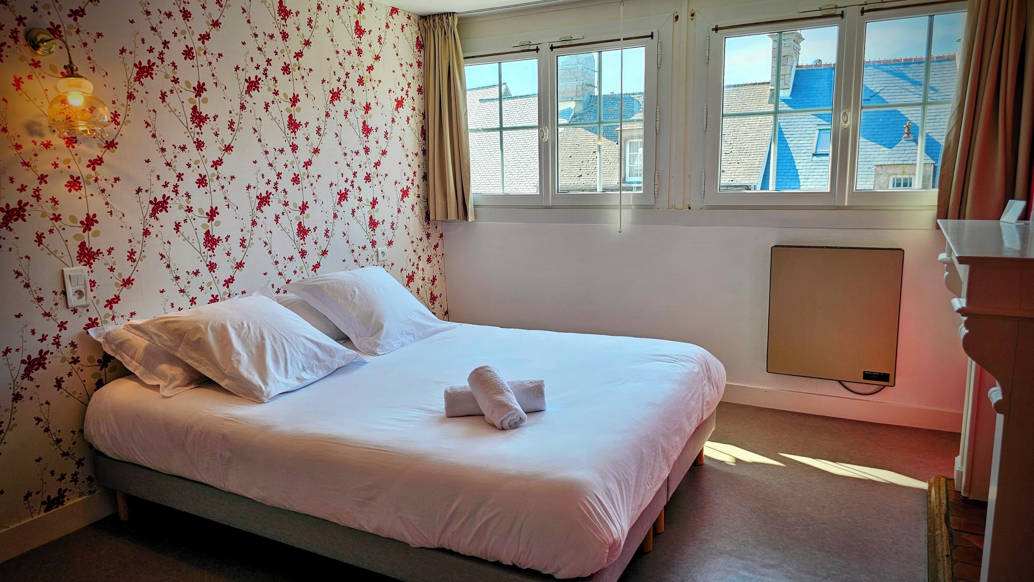 Habitación de hotel con cama, papel tapiz floral, ventana con vistas a edificios y un radiador.