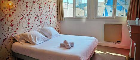 Habitación de hotel con cama, papel tapiz floral, ventana con vistas a edificios y un radiador.