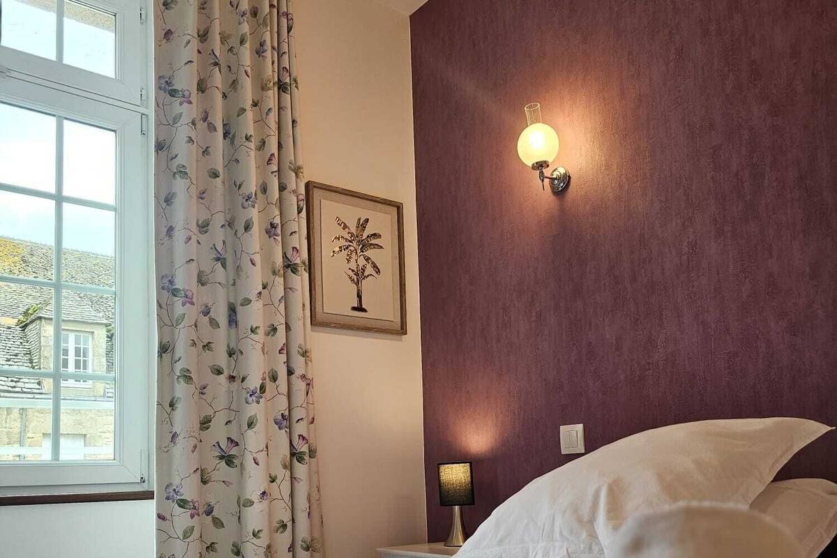 Une chambre d’hôtel avec un lit, une table de chevet, une lampe, une fenêtre avec des rideaux et un tableau encadré au mur.
