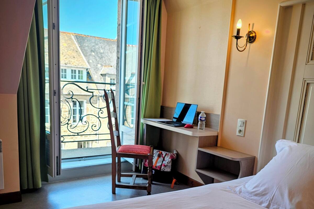 Une chambre d’hôtel avec un lit, un bureau avec un ordinateur portable, une chaise, une fenêtre avec des rideaux et une vue sur les bâtiments environnants.