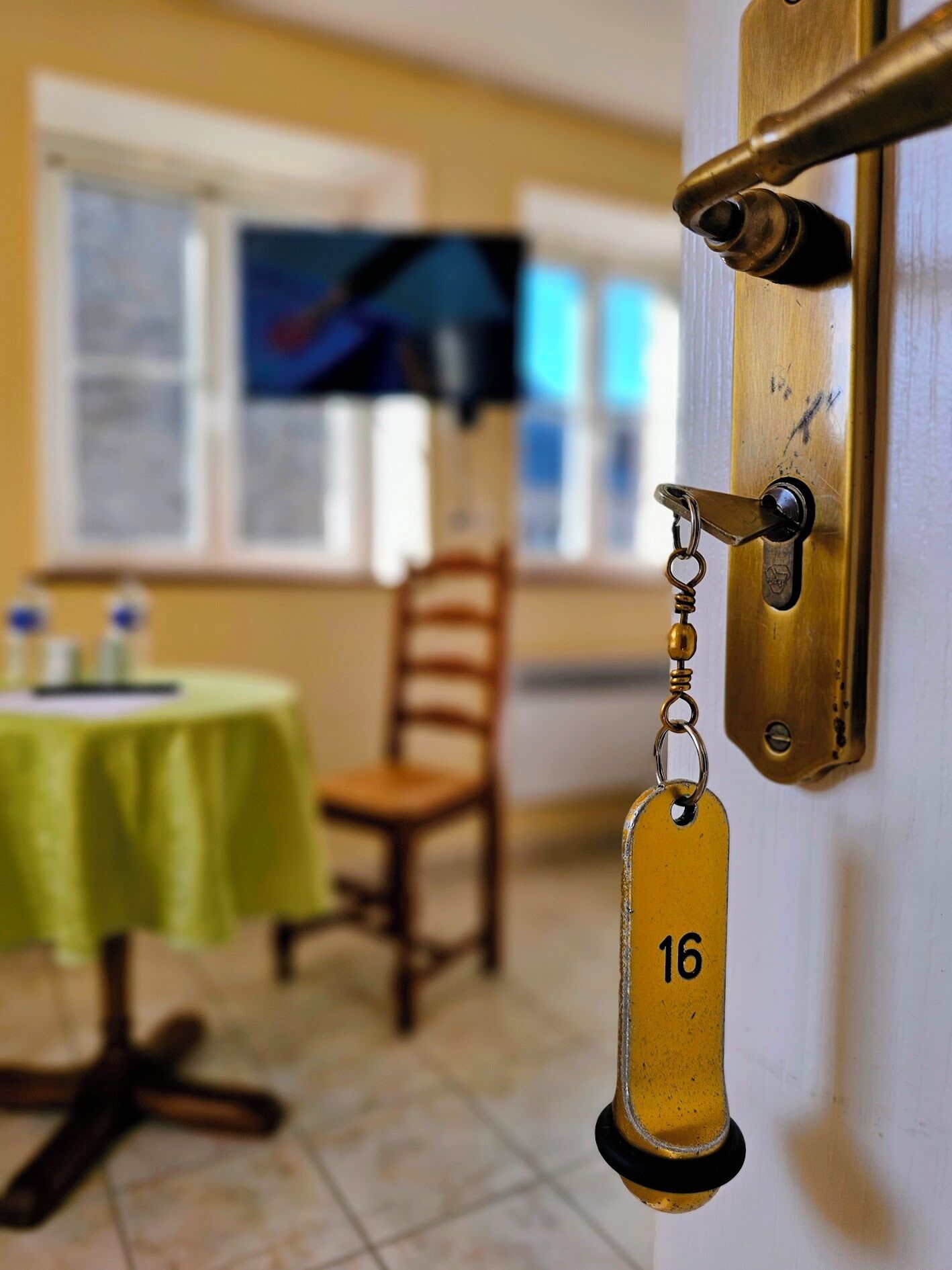 Puerta de habitación de hotel con picaporte de latón, llavero con el número 16, una mesa verde y una silla de madera.