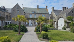 A stone building with a central entrance, surrounded by a gravel path and greenery. - Le Conquérant (Barfleur)