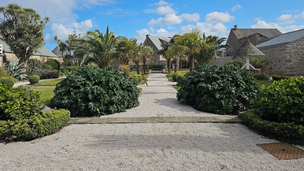 A gravel pathway leading to a stone building with a thatched roof, surrounded by lush greenery and palm trees. - Le Conquérant (Barfleur)