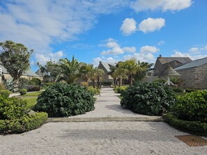 Un chemin de gravier mène à un bâtiment en pierre surmonté d’un toit de chaume, entouré d’une végétation luxuriante et de palmiers.