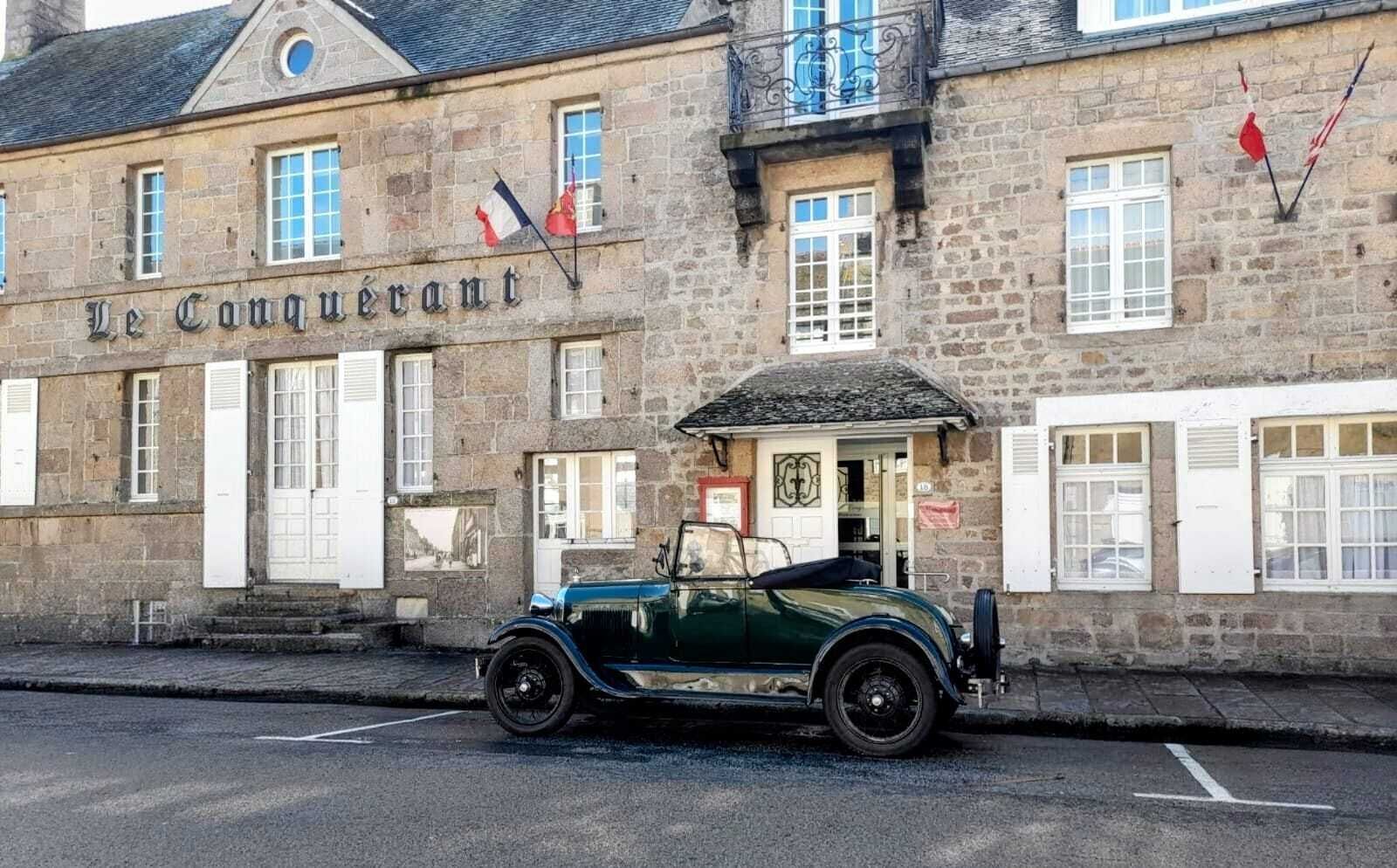 A stone building with a sign reading "Le Conquérant" and a vintage green car parked in front.