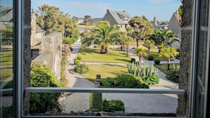 A view through a window of a courtyard with palm trees, a stone bridge, and a stone building in the background. - Le Conquérant (Barfleur)