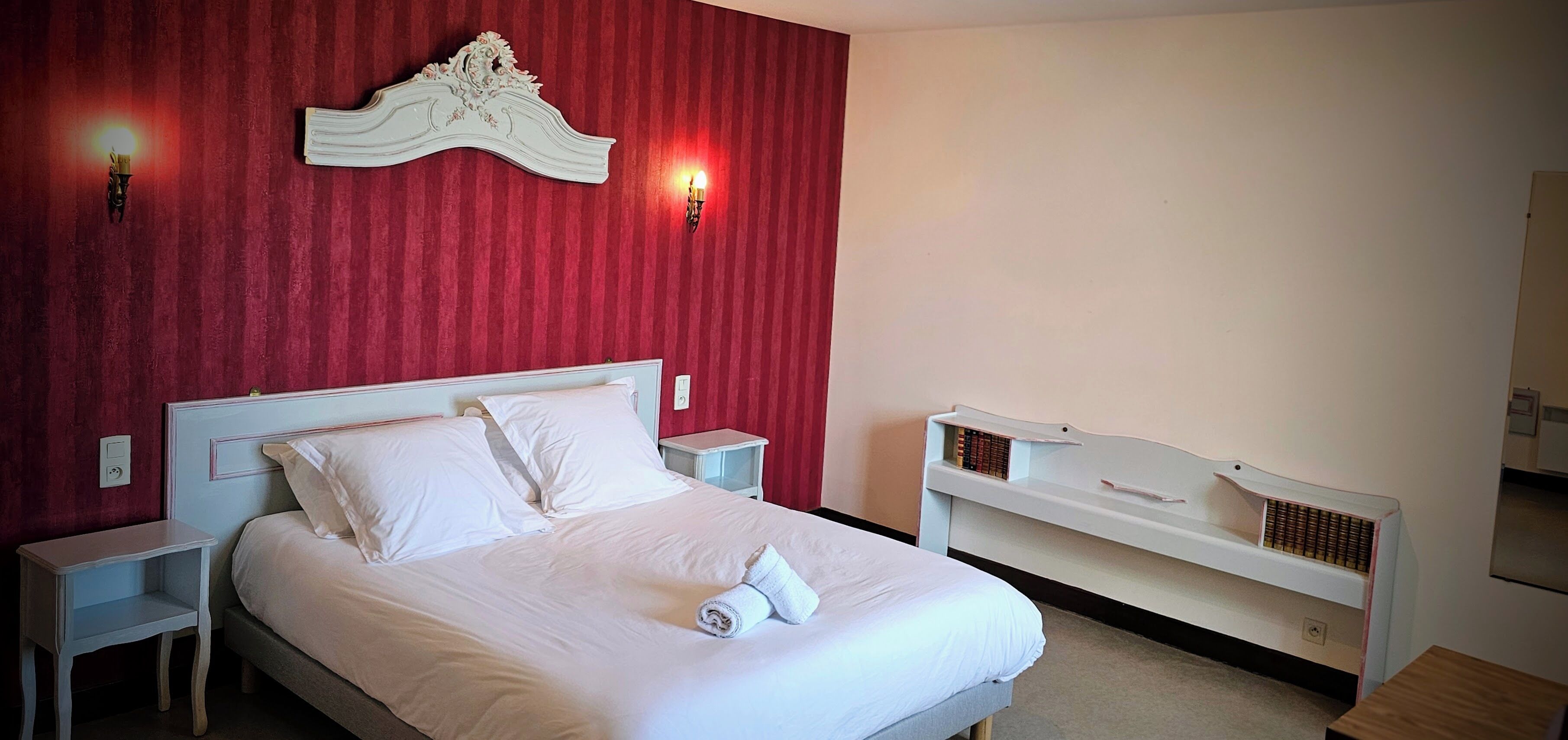 Un lit bien fait, avec du linge de lit blanc et des oreillers, une table de chevet, une commode, une tête de lit blanche et une petite étagère près du lit avec des livres.