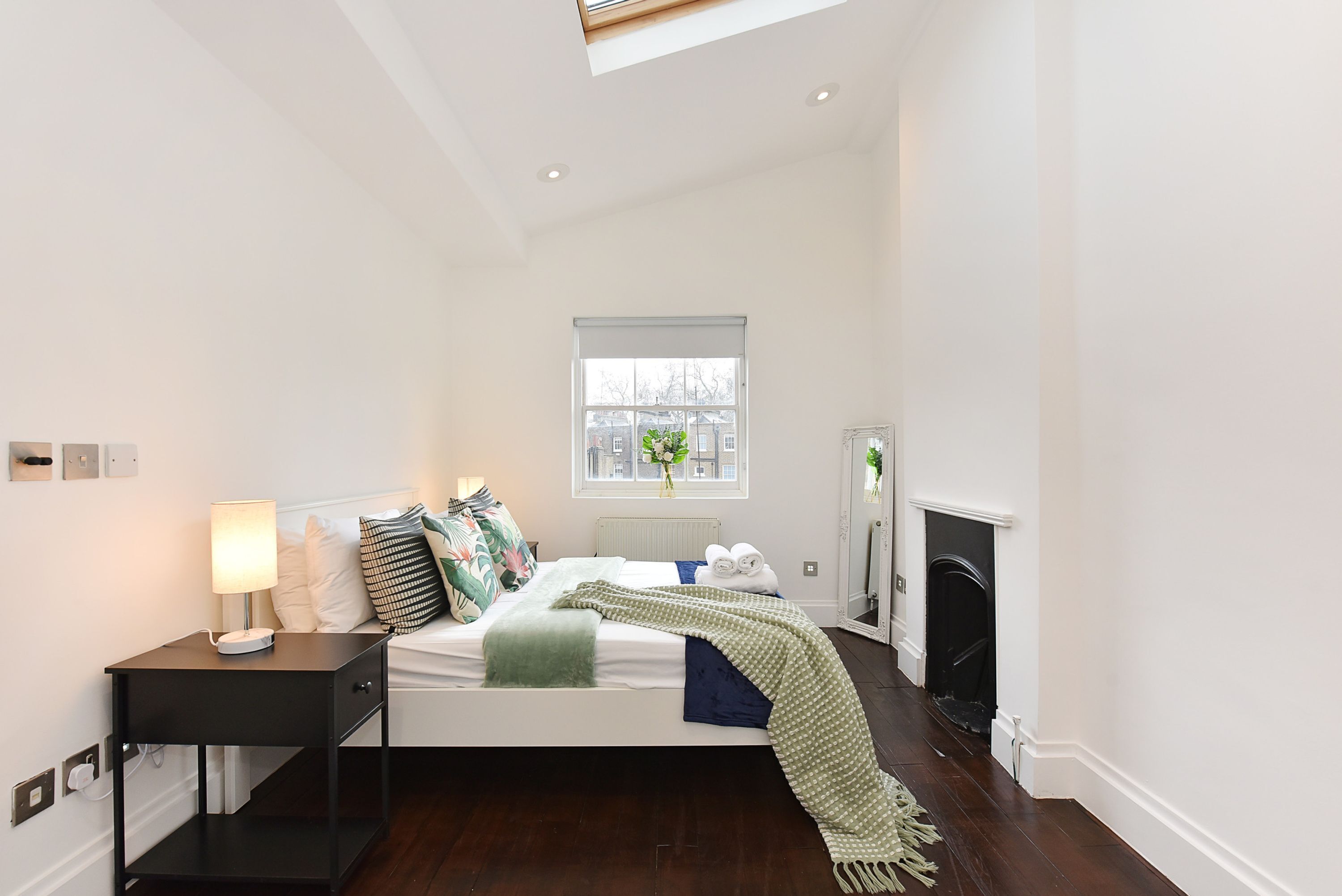A bedroom with a bed, bedside table, lamp, window, fireplace, and a view of the outside.