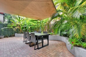 Un espace patio aménagé avec une table et des chaises, entouré d’une végétation luxuriante et agrémenté d’un jardinier en béton aux formes arrondies.
