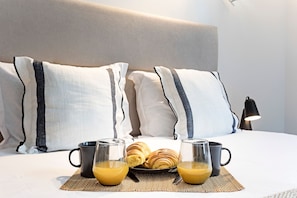 A neatly made bed with pillows, a plate of croissants, and two glasses of orange juice.