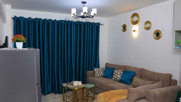 Ett kompakt bostadsutrymme med en soffa, ett litet runt bord, ett kylskåp och en gardin.