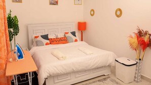 En snyggt bäddad säng med vita sängkläder och dekorativa kuddar, ett litet sängbord med ett strykjärn, en lampa och en papperskorg.