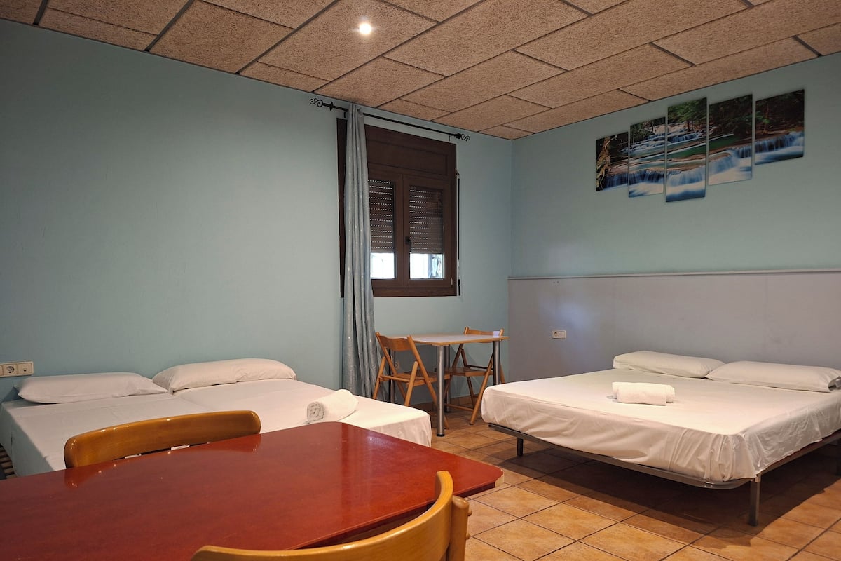 A room with two beds, a wooden table, a chair, a window with curtains, and a wall-mounted picture.