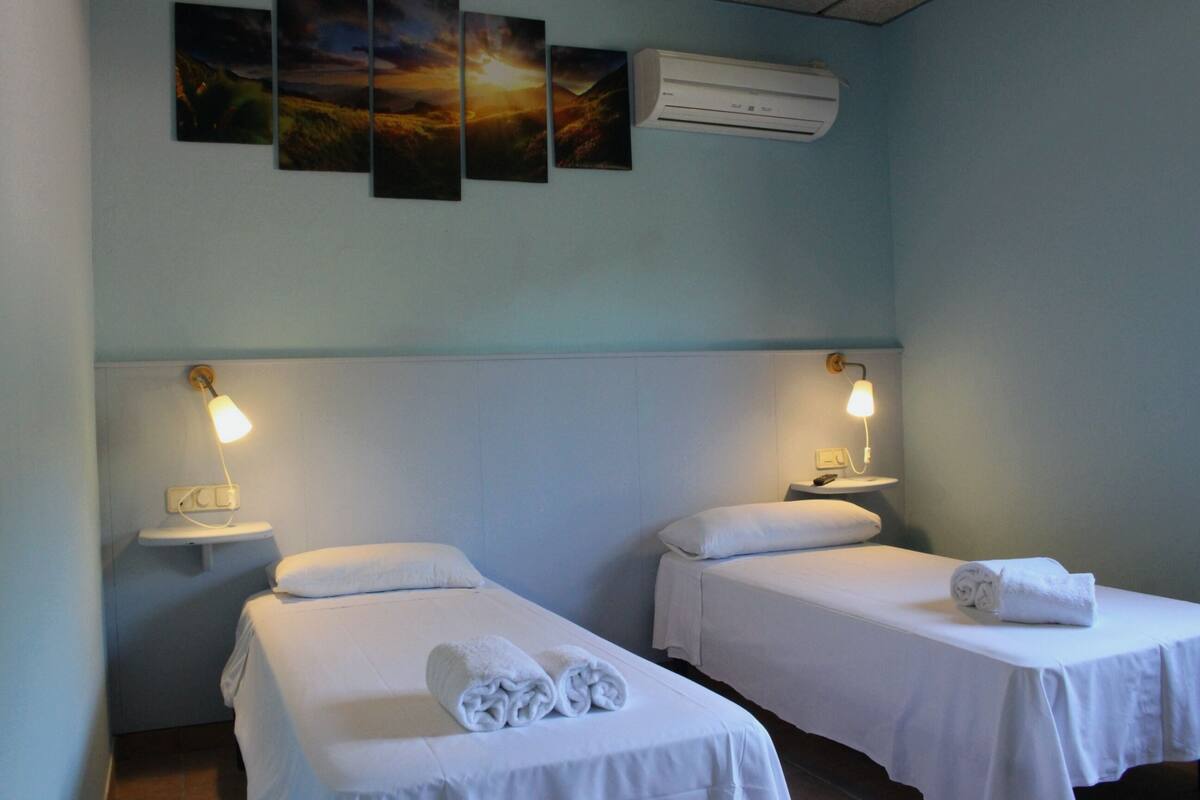 Two single beds with white linens, a wall-mounted air conditioner, and a nightstand with a lamp.