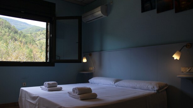 A bedroom with a bed, two bedside lamps, a window with a view of a forest, and an air conditioning unit. - Hotel Al Riu  (Bagà)