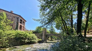 A stone bridge over a clear stream, surrounded by lush greenery and a historic stone building. - Hotel Al Riu  (Bagà)
