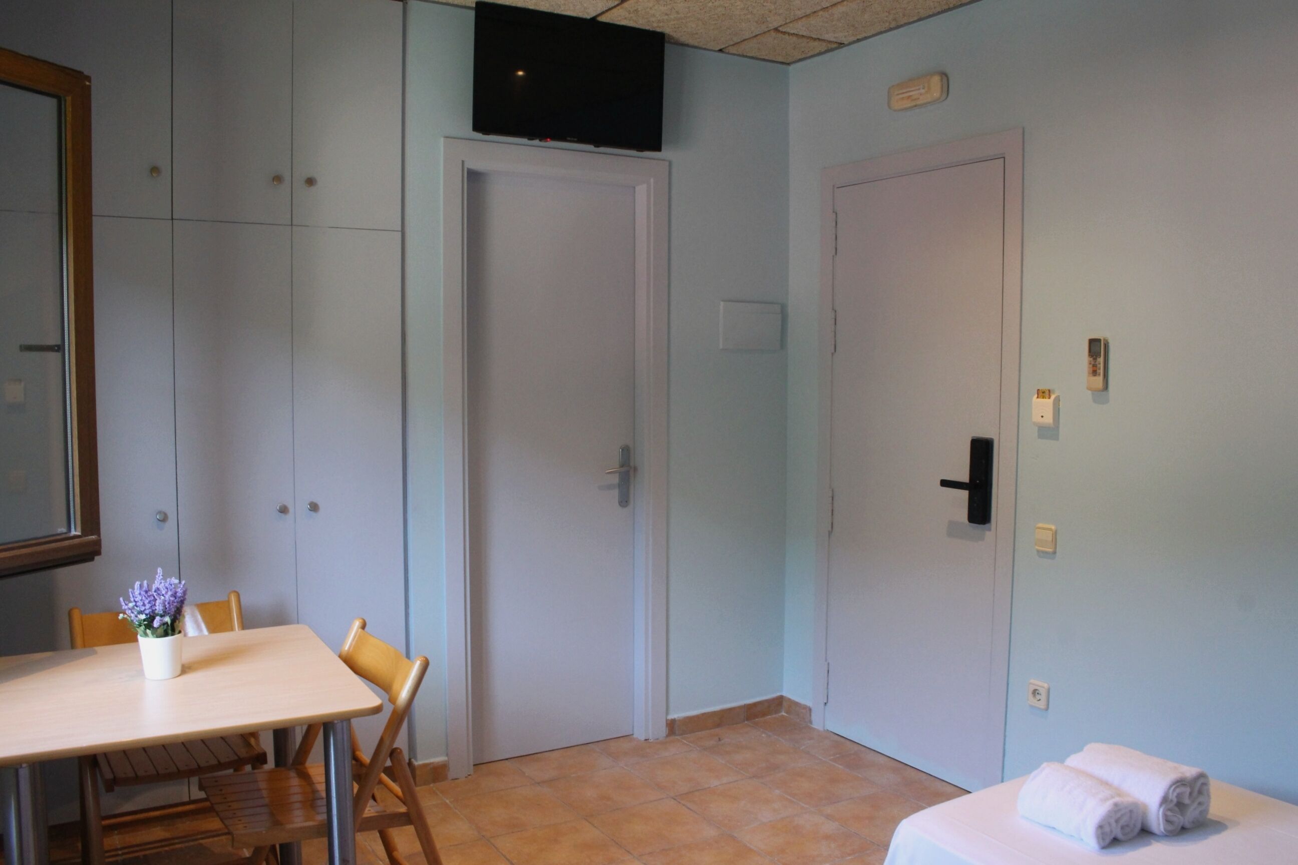A room with a bed, a table with chairs, a television mounted on the wall, and a door.