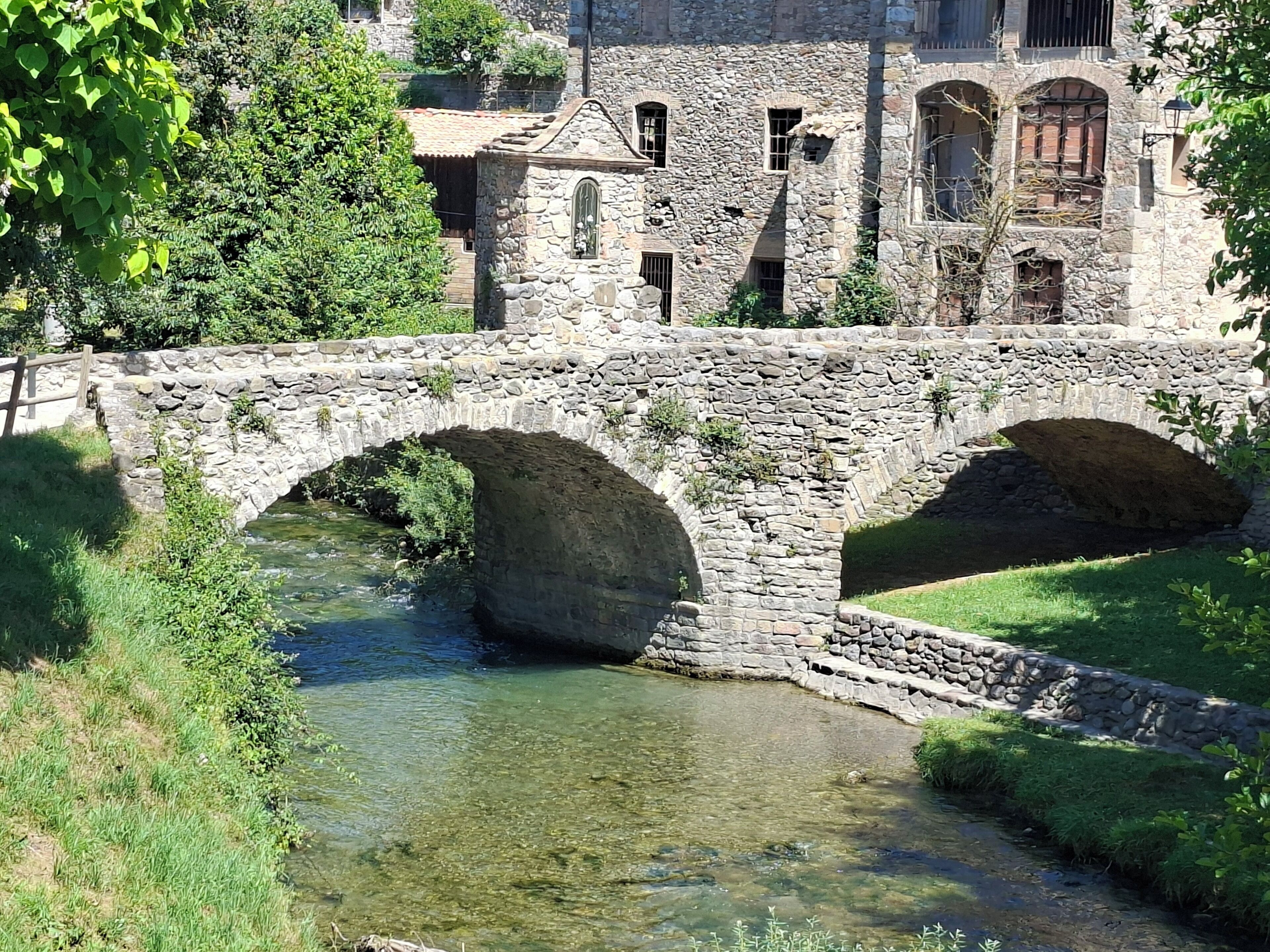 A stone bridge over a clear stream with a stone building in the background.