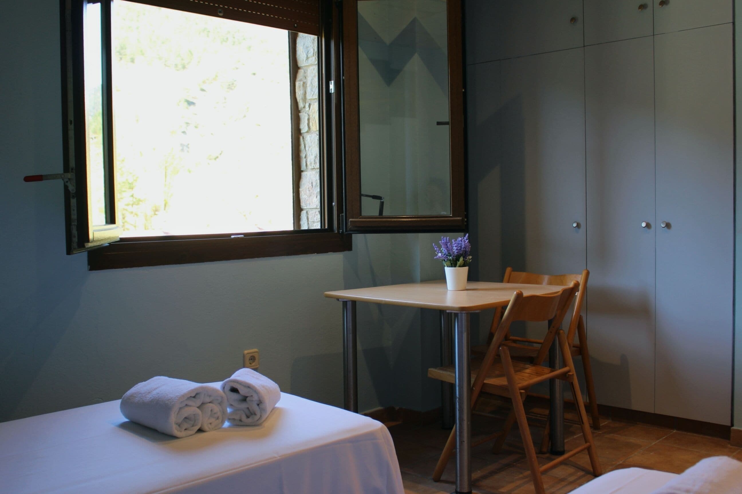 Una habitación con una cama, una mesita con una silla, una ventana con vistas a árboles y un armario.