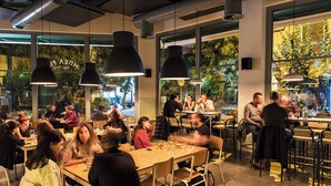 Ein modernes Restaurant mit großen Fenstern, mehreren Esstischen und hängenden Pendelleuchten.