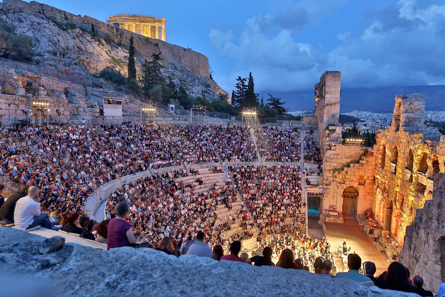 A large outdoor amphitheater with tiered seating and a stage, surrounded by ancient ruins and a rocky hillside.