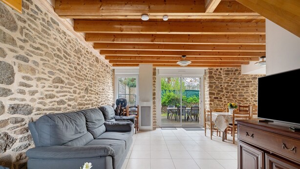 A living room with a stone wall, wooden ceiling beams, a gray sofa, a TV on a wooden cabinet, and a view through a glass door to an outdoor patio.
