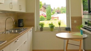 Een kleine keuken met raam, een spoelbak, kastjes, een koffiezetapparaat en een tafeltje.
