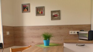 Een kleine keuken met een houten tafel, een blauwe plant in pot, een broodrooster en een muur met drie ingekaderde foto's.