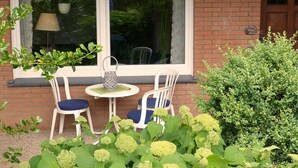 Een bakstenen gebouw met een raam, twee witte stoelen met blauwe kussens, een klein rond tafel en een bloempot met plant.
