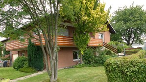 Een huis met twee verdiepingen, een houten terras, een voortuin met een boom en een haag.