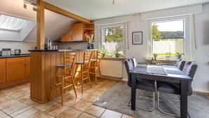 Een moderne keuken met houten kastjes, een eethoek met een tafel en stoelen, en uitzicht op groen buiten.