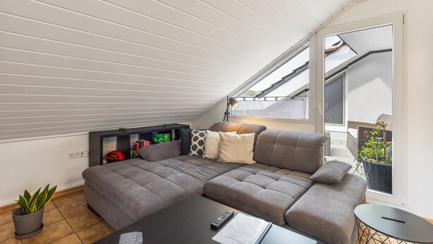 Een moderne woonkamer met een modulaire bank, een salontafel en een bijzettafeltje. Er is een boekenplank met boeken en een spel, en een potplant. De kamer heeft een schuine nok met dakramen.