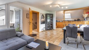 Een moderne woonkamer met een grijze bank, een eethoek met een tafel en stoelen, een keuken met houten kastjes en een gang die leidt naar een trap.