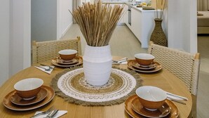 Een eethoek met een ronde houten tafel voor vier personen, omringd door rieten stoelen. In het midden staat een witte vaas met gedroogde rietstengels. De kamer heeft een neutrale kleurenpalet met witte muren en een houten plafond.