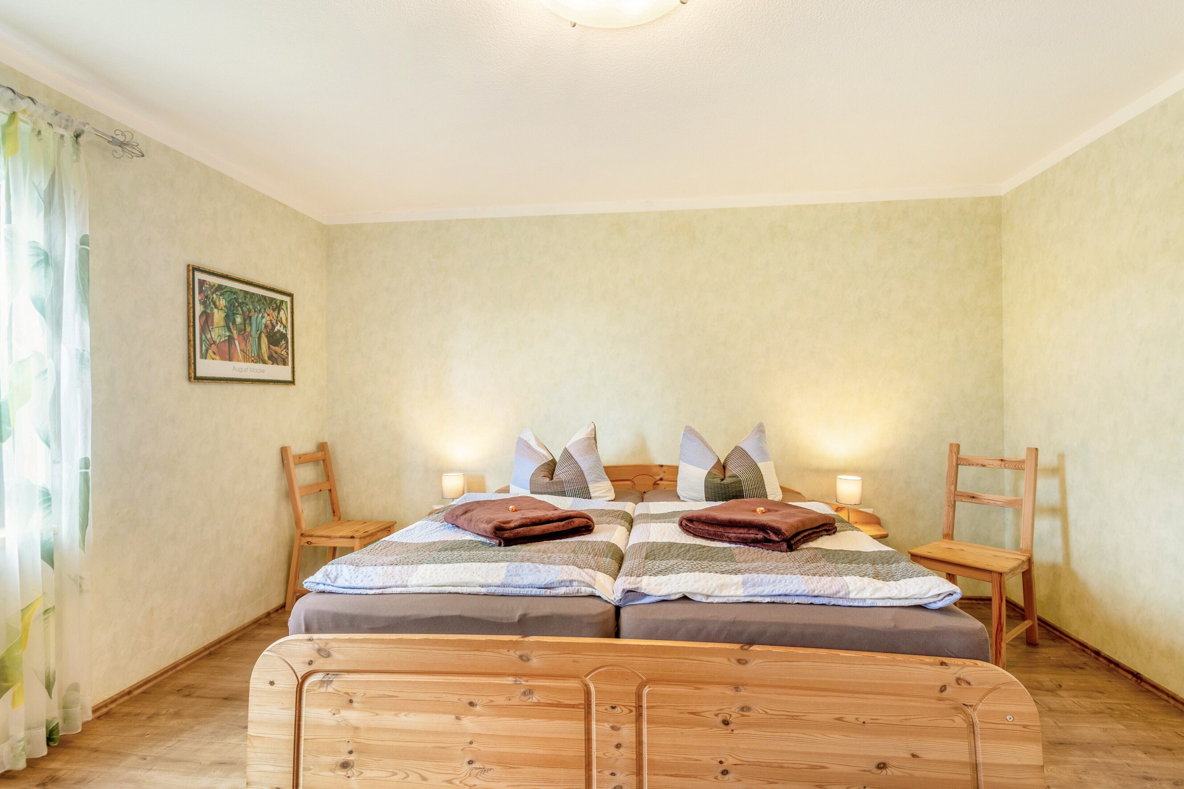 A double bed with a wooden headboard, two bedside tables with lamps, a wooden chair, and a painting on the wall.