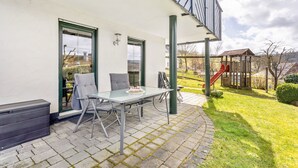 A patio with a table and chairs, a trash bin, and a playground in the background. - Apartment "64 sqm" with Private Terrace, Shared Garden, and Wi-Fi (Olsberg)