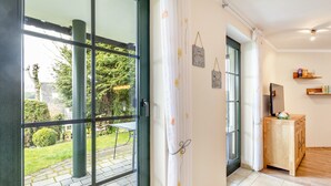 A room with a sliding glass door leading to a garden, a wooden cabinet, and a wall-mounted shelf with items. - Apartment "64 sqm" with Private Terrace, Shared Garden, and Wi-Fi (Olsberg)