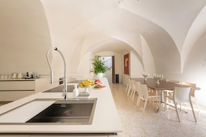A modern kitchen with a central island, a dining area with a wooden table and chairs, and a view of a staircase leading to an upper level. - Garden Apartment - Palazzo Villani Lubelli (Cannole)