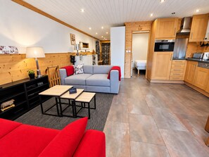 Un salon moderne avec un canapé gris, des coussins rouges, une table à manger en bois et un coin cuisine doté de meubles et d’appareils électroménagers en bois.