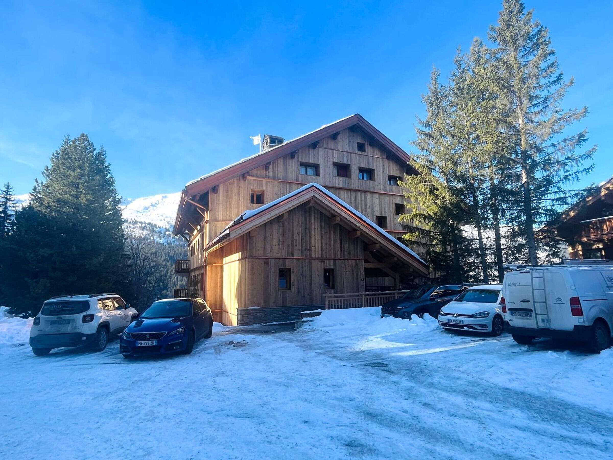 Una cabaña de madera con techo inclinado, rodeada de nieve y coches aparcados.