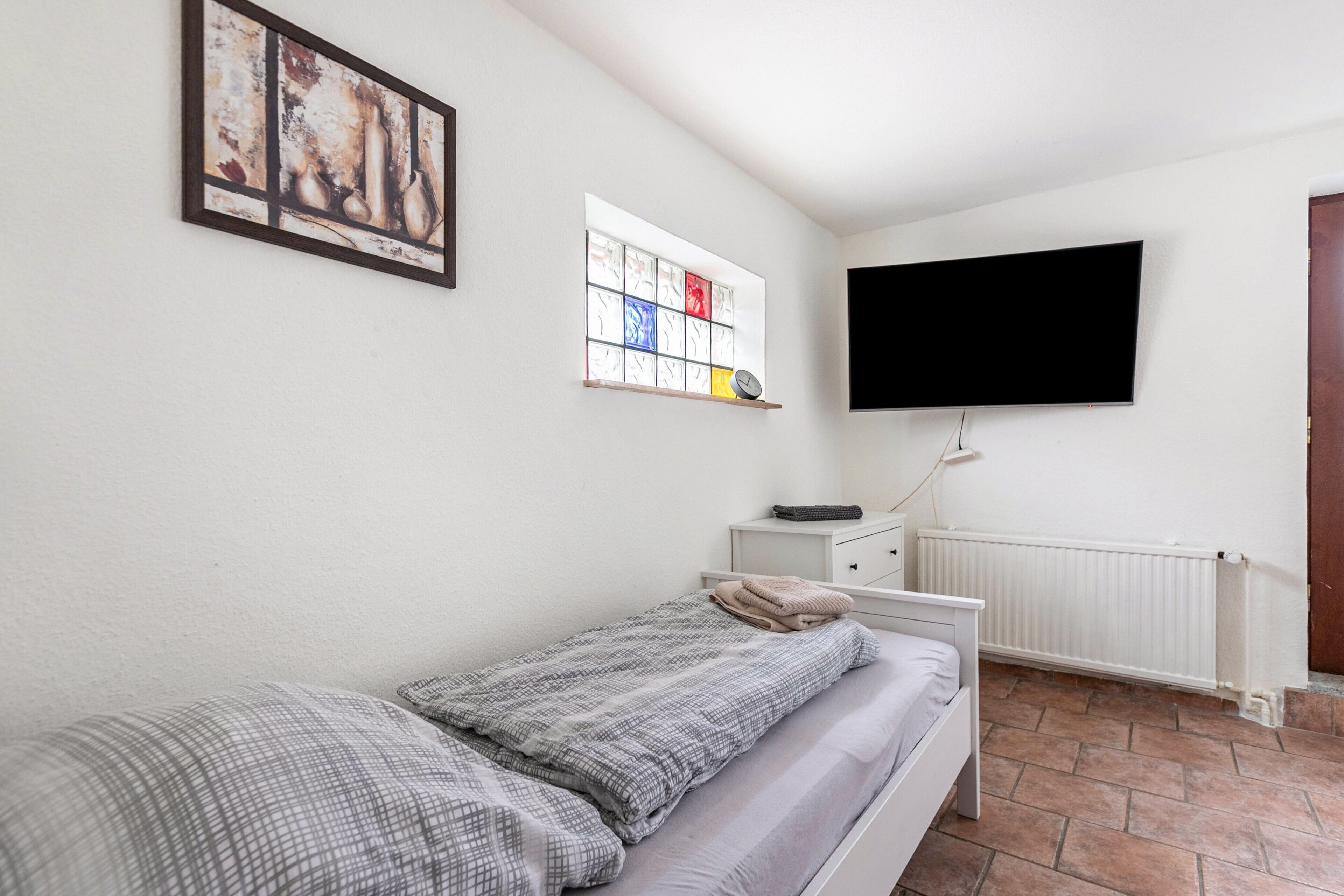 Een slaapkamer met een bed, een aan de muur bevestigde televisie, een raam met een glas-in-lood ontwerp en een radiator.