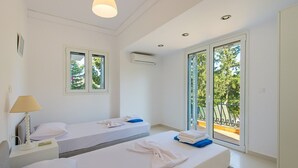 Ein Hotelzimmer mit zwei Betten, einem Nachttisch, einer Lampe und einem Balkon mit Blick auf Bäume.