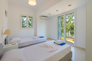 A hotel room with two beds, a nightstand, a lamp, and a balcony with a view of trees. - Giovanni · Villa Giovanni (Faliraki)