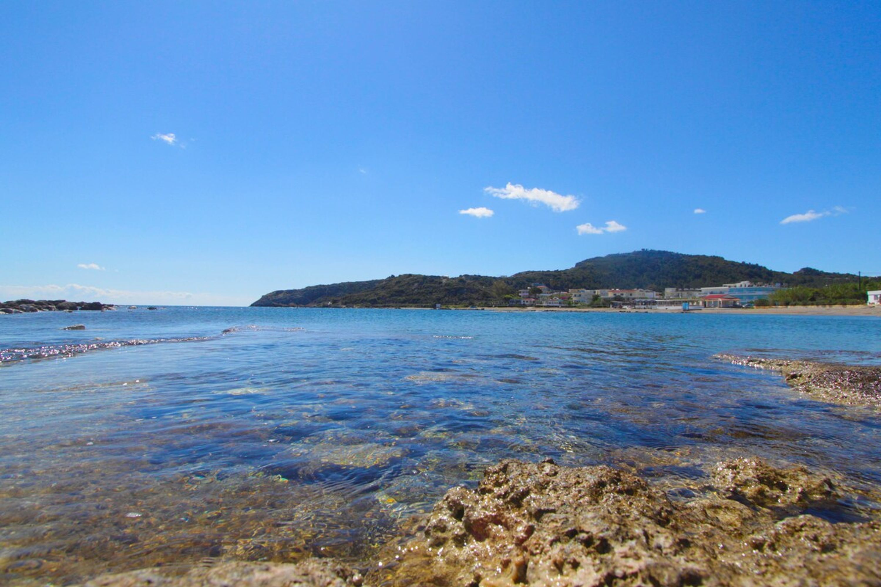 A coastal scene with clear water, rocky shore, and a distant hill with buildings.