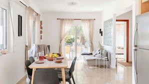 A modern living room with a sofa, a dining table with chairs, and a kitchen area with a refrigerator. - Villa Katherina - Tsambika View (Monastiri i Tsampika)