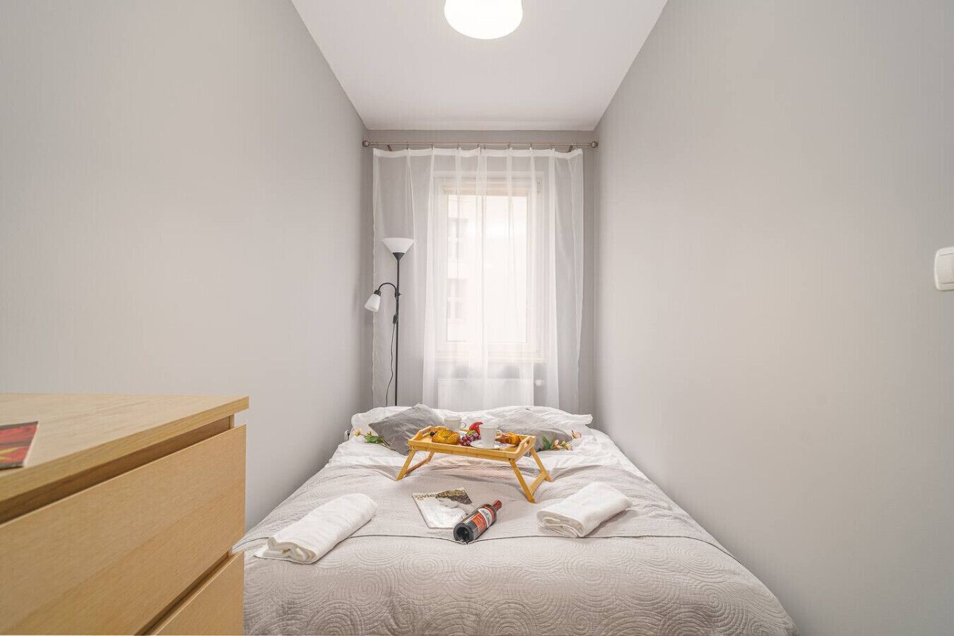 A small, neatly arranged bedroom with a single bed, a bedside table, and a window with sheer curtains.