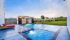 A modern outdoor swimming pool with a geometric design, a lounge area with red cushions, and a view of a landscaped garden and distant hills. - Delight The Lavish Loft Corbett (Kaladhungi)