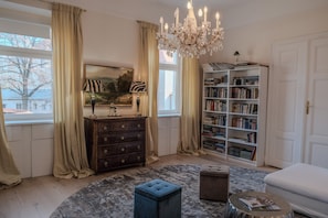 Ein Zimmer mit großem Fenster, einer Kandelaberleuchte, einem Bücherregal, einem Schmuckkästchen und einem runden Teppich.