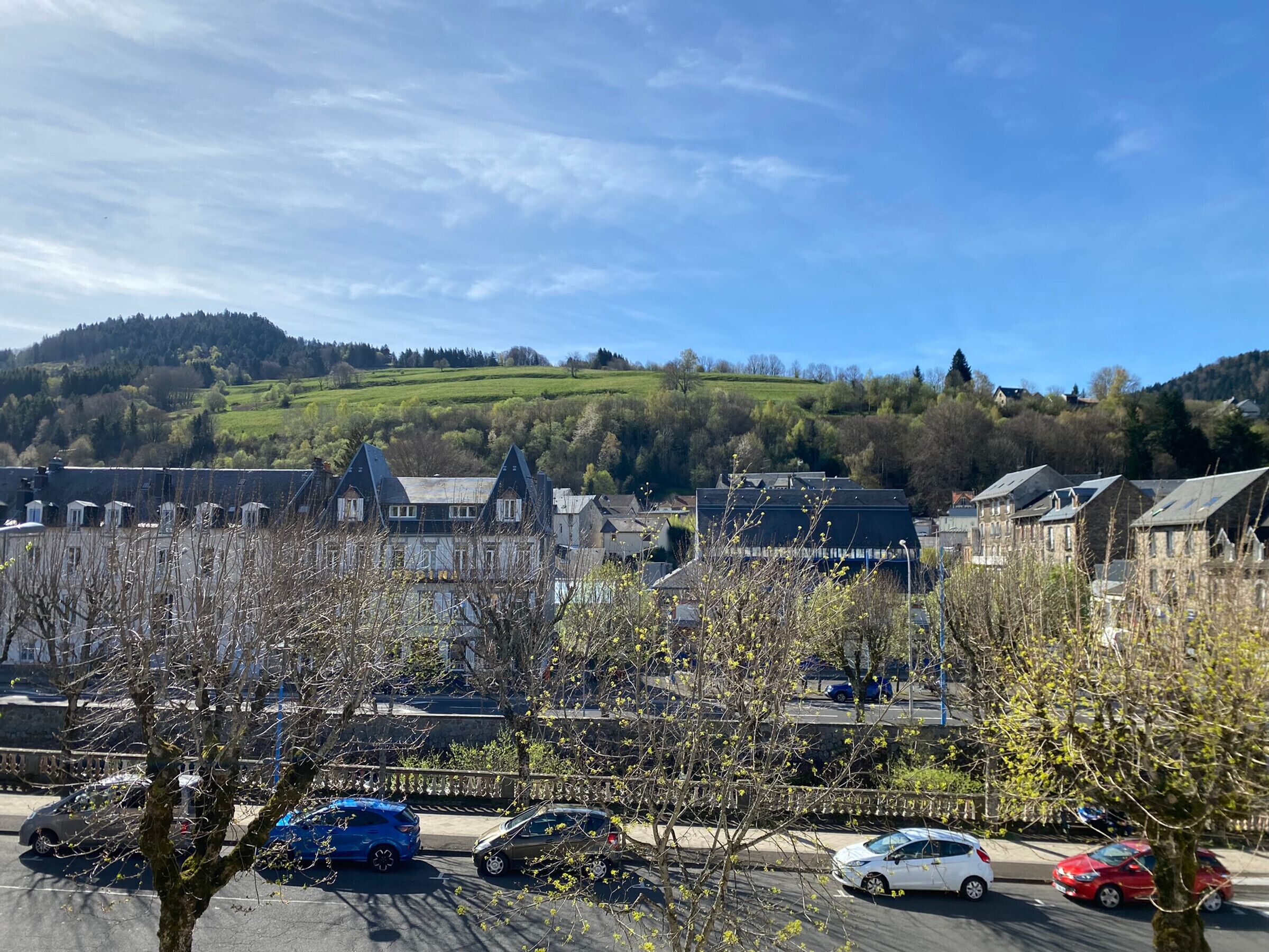 Un parking où sont garées plusieurs voitures, entouré de bâtiments à l’architecture traditionnelle, avec un paysage vallonné en arrière-plan.